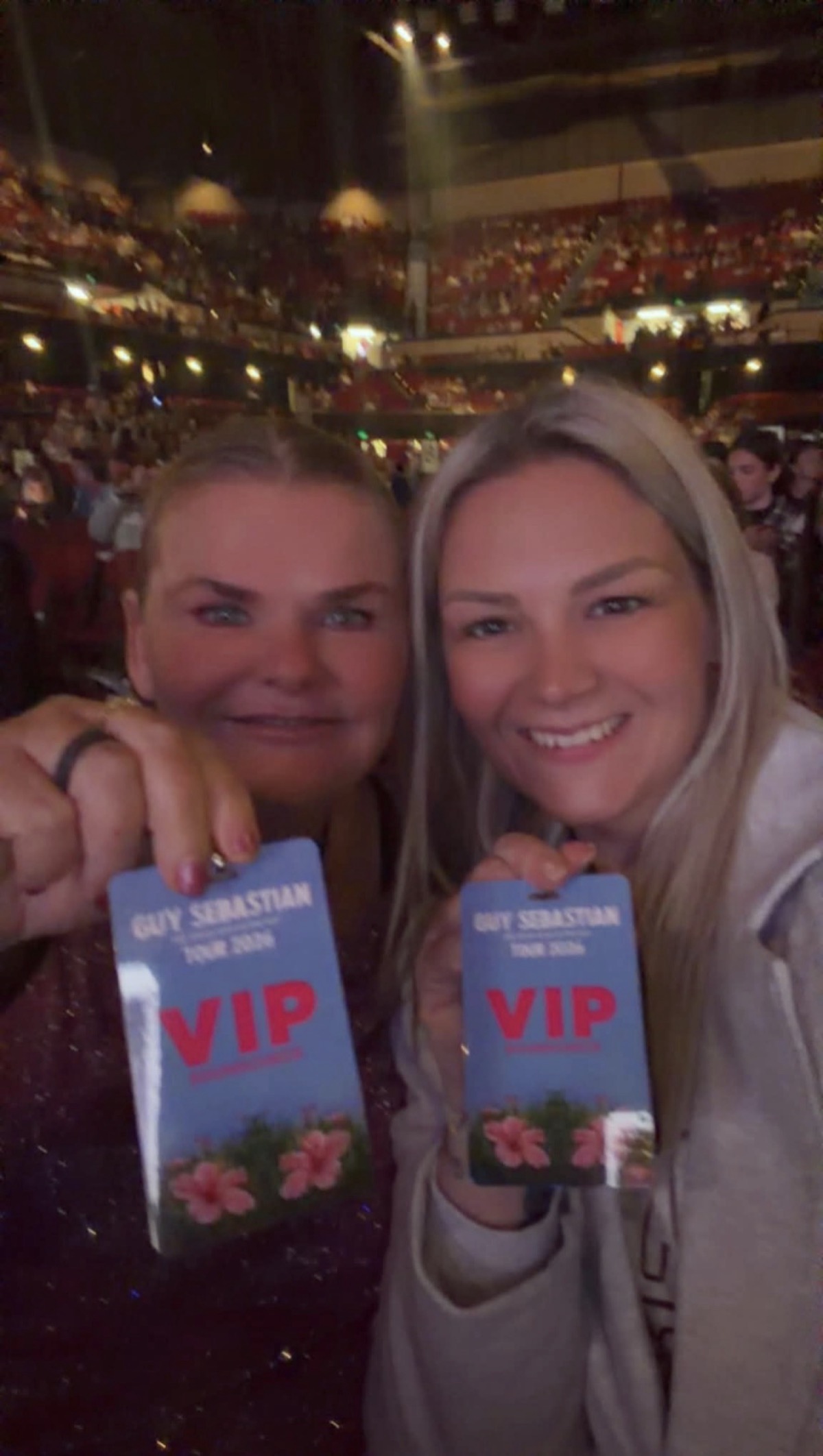 Two friends together at a Guy Sebastian concert, smiling and holding up their VIP passes