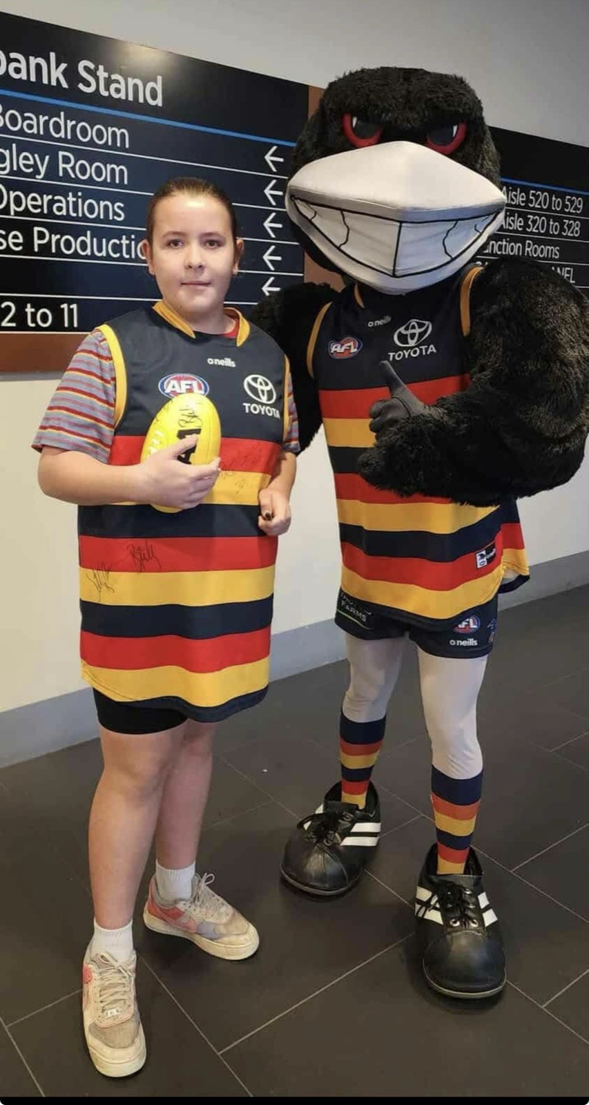 A big smile in a team guernsey next to the club mascot at the stadium, holding a signed football